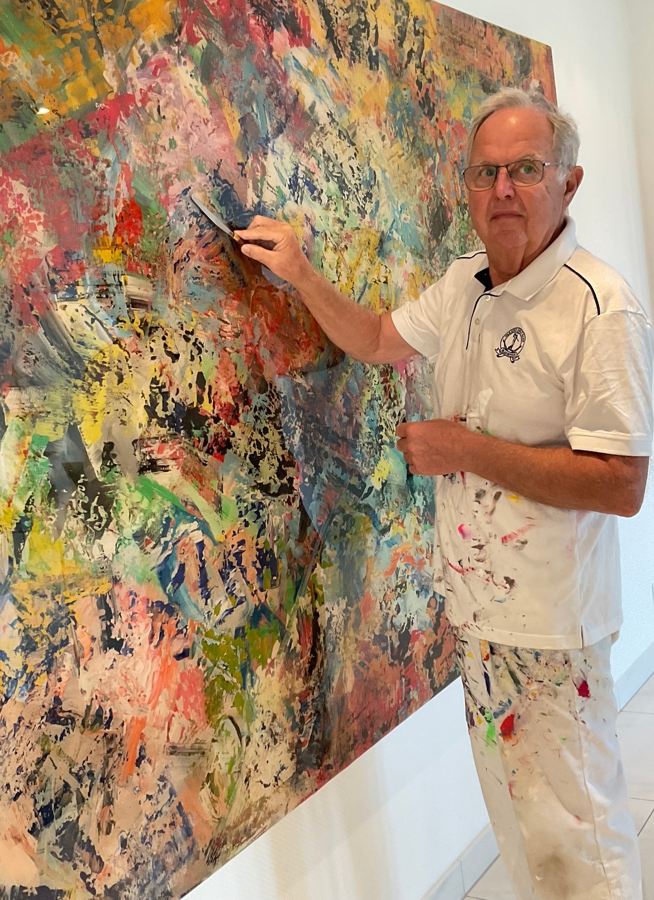 Ein älterer Mann mit Brille in einem farbverschmierten Hemd und Hose malt ein abstraktes Gemälde mit einem Spachtel.
